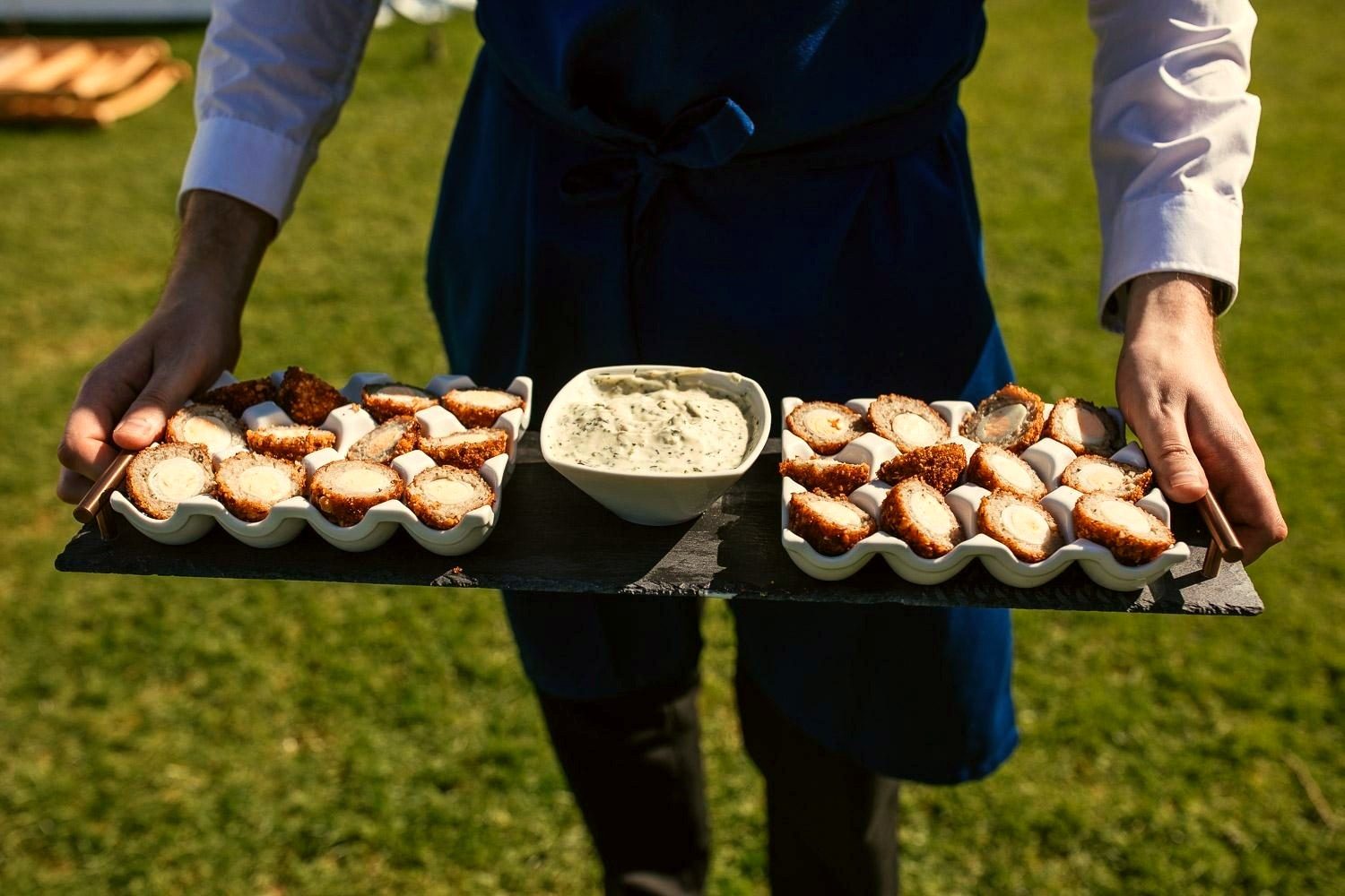 A waiter presenting a tray of scotch egg canapes with herb dip.