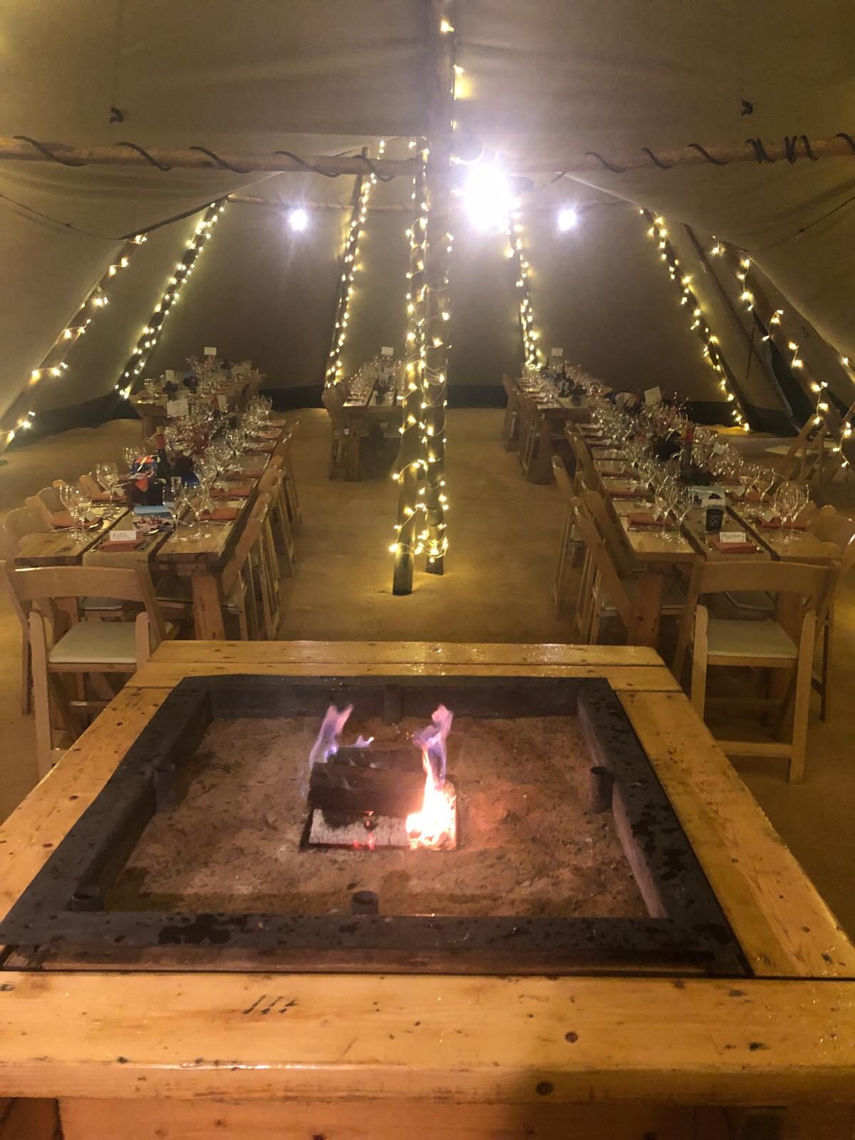 A tipi laid for dinner with a central fire pit and fairy lights.